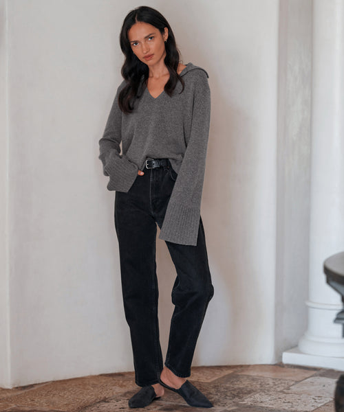 A woman with long dark hair wears the Cashmere Frances Polo in grey, paired with black jeans and black flats. She stands indoors against a light wall, looking at the camera with a neutral expression.