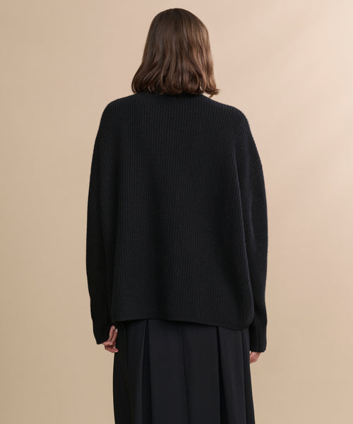 A person with shoulder-length brown hair faces away, wearing the Cashmere Half Zip in black and a long black skirt, set against a neutral beige background.