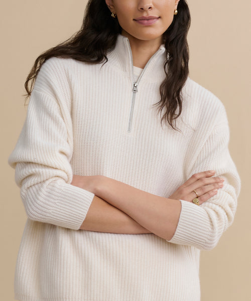 A woman with long brown hair wears the Cashmere Half Zip sweater with sleeves rolled up, standing with her arms crossed against a beige background.