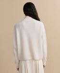 A person with long dark hair is shown from behind, wearing the Cashmere Half Zip in ivory and a matching cream skirt, standing against a beige background.