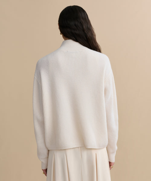 A person with long dark hair is shown from behind, wearing the Cashmere Half Zip in ivory and a matching cream skirt, standing against a beige background.