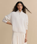 A woman with long, wavy dark hair wears the Cashmere Half Zip sweater in ivory with a matching pleated skirt, standing against a neutral beige background.