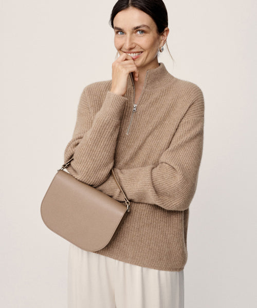 A woman with dark hair tied back smiles while wearing the Cashmere Half Zip in undyed stone, paired with cream pants and holding a taupe handbag under her arm against a plain light background.
