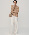 A woman stands against a plain background, smiling with one hand partly covering her mouth. She wears the undyed stone Cashmere Half Zip sweater, white wide-leg pants, open-toed shoes, and carries a beige shoulder bag.