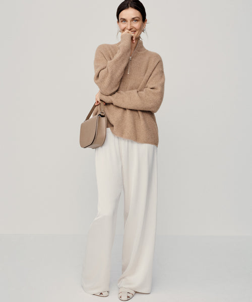 A woman stands against a plain background, smiling with one hand partly covering her mouth. She wears the undyed stone Cashmere Half Zip sweater, white wide-leg pants, open-toed shoes, and carries a beige shoulder bag.