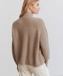 A woman with light brown hair is seen from the back, turned slightly to the side, wearing the Cashmere Half Zip in undyed stone with white pants against a plain light background.