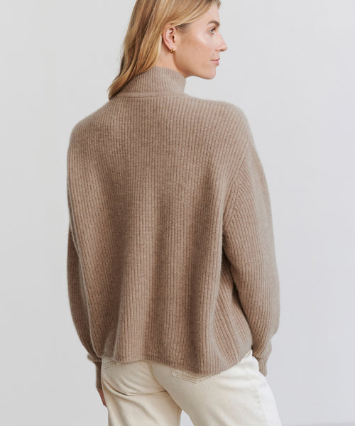 A woman with light brown hair is seen from the back, turned slightly to the side, wearing the Cashmere Half Zip in undyed stone with white pants against a plain light background.