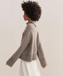 A woman with curly hair is shown in profile wearing the Cashmere Jackie Sweater—a luxe taupe knit with ribbed cuffs and hem—paired with a flowing white skirt, standing against a neutral background.