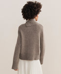 A person with curly hair, seen from behind, wears the chunky taupe Cashmere Jackie Sweater with button-shoulder detail and a white pleated skirt, standing against a plain light background.