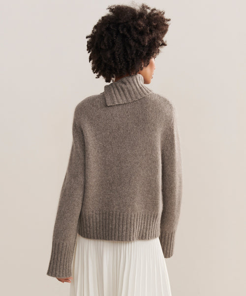 A person with curly hair, seen from behind, wears the chunky taupe Cashmere Jackie Sweater with button-shoulder detail and a white pleated skirt, standing against a plain light background.