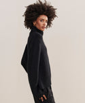 A woman with voluminous curly hair models the Cashmere Jackie Sweater, a luxury black knit turtleneck, paired with black pants. She stands sideways against a plain light background, looking at the camera.