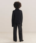 A person with curly hair, seen from behind, is wearing the Cashmere Jackie Sweater in black with loose black pants, standing on a light-colored carpet against a plain neutral wall.
