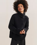 A woman with natural curly hair wears the Cashmere Jackie Sweater, a luxury black knit turtleneck with button-shoulder detailing, paired with black pants against a plain light background.