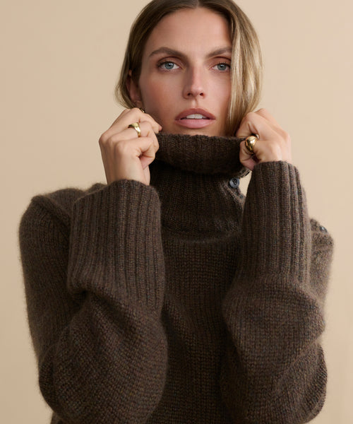 A person with long hair wears the Cashmere Jackie Sweater in brown, holding the collar up to their chin against a beige background. They face the camera directly with a neutral expression.