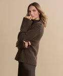 A woman with light brown hair stands sideways against a beige background wearing the Cashmere Jackie Sweater—a luxe knit brown top with button-shoulder detailing—and a dark skirt, gently touching her lips with one hand and pointing with the other.