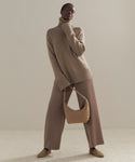 Wearing the Cashmere Jet Turtleneck, wide-legged mauve pants, and beige shoes, a person stands holding a matching beige handbag against a neutral background.