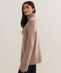 A woman with long dark hair models the Cashmere Jet Turtleneck, featuring ribbed cuffs and hem, as she stands sideways and looks at the camera against a plain light background.