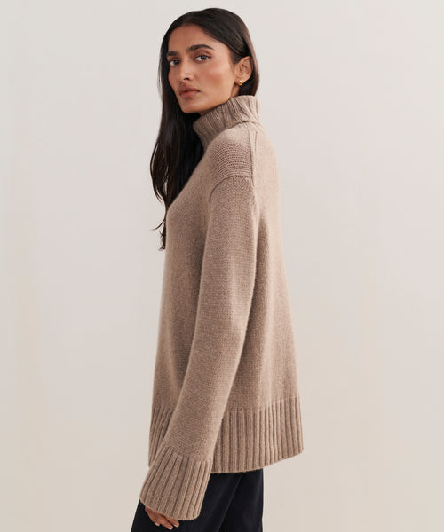 A woman with long dark hair models the Cashmere Jet Turtleneck, featuring ribbed cuffs and hem, as she stands sideways and looks at the camera against a plain light background.