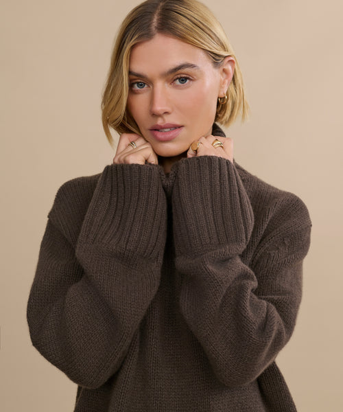 A woman with blonde hair wears the Cashmere Jet Turtleneck, holding the collar with both hands. She looks at the camera with a neutral expression, accessorized with gold rings and small hoop earrings against a beige background.