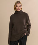 A woman with short blonde hair wears the Cashmere Jet Turtleneck and black pants, standing against a plain beige background and looking toward the camera with a neutral expression.