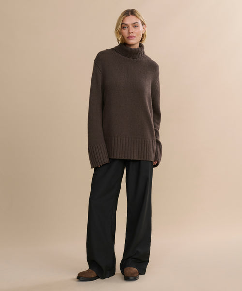 A person with short blonde hair and a relaxed expression poses against a beige background, wearing the oversized Cashmere Jet Turtleneck with wide-leg black pants and brown shoes.