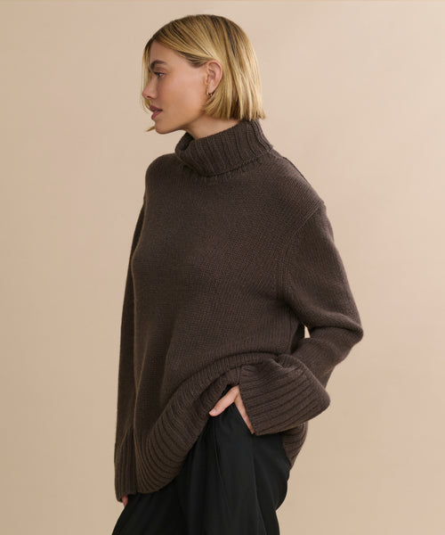 A person with a short blond bob hairstyle wears the loose, oversized Cashmere Jet Turtleneck and black pants, standing sideways against a plain beige background.