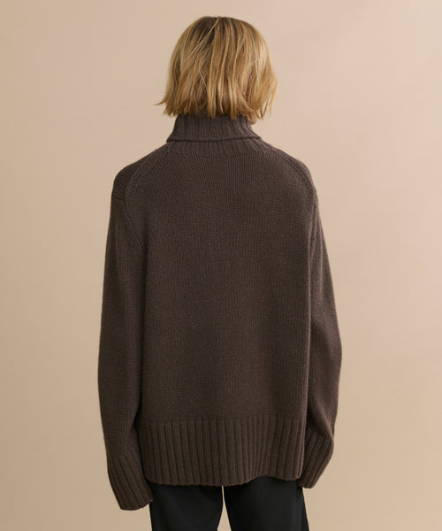 A person with blonde, chin-length hair is shown from behind wearing the Cashmere Jet Turtleneck in dark brown and dark pants against a plain beige background.