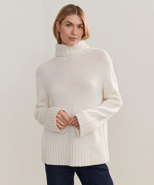 A woman in the cream Cashmere Jet Turtleneck and dark jeans stands against a light background, looking at the camera with a soft smile and relaxed posture.