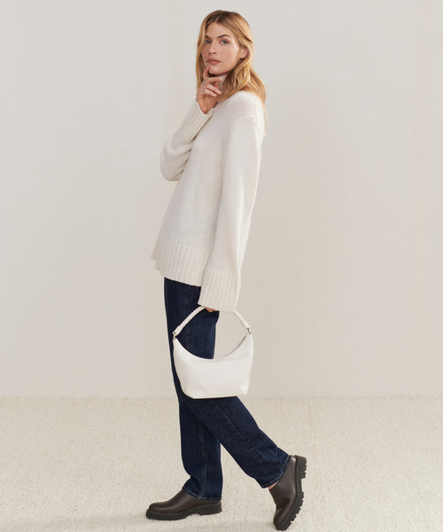 A woman stands sideways against a plain light background, wearing the Cashmere Jet Turtleneck with dark blue jeans and black boots. She holds a small white handbag and gazes forward, one hand near her chin.