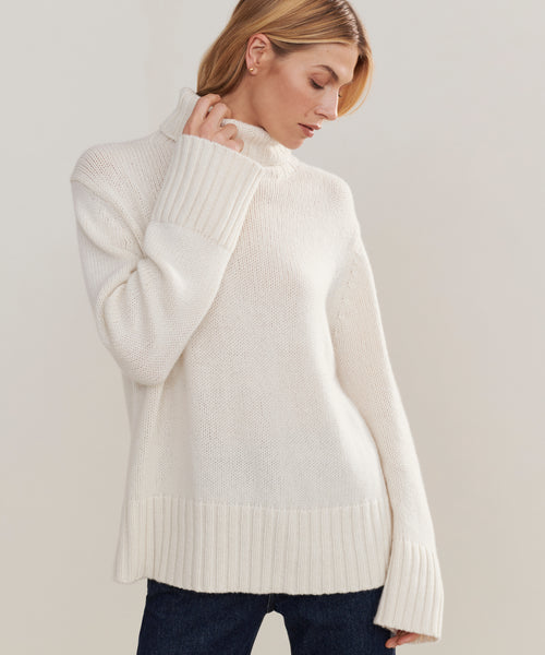 Someone wears the Cashmere Jet Turtleneck with long sleeves and dark jeans, standing against a plain background, looking down, and touching the collar.