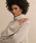 A woman with curly hair models the beige Cashmere Jet Turtleneck, posing confidently against a neutral background with one hand resting on her shoulder.
