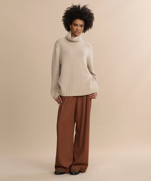 A person with curly hair models the oversized Cashmere Jet Turtleneck in beige with loose brown trousers, standing against a plain beige background.