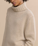 A person wears the Cashmere Jet Turtleneck, a chunky, oversized sweater in light beige, standing against a plain beige background. The image is cropped at the mouth and above the waist.