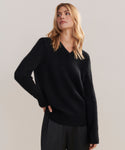 A woman with long blonde hair models the Cashmere Kate V-Neck sweater in black, featuring a relaxed boyfriend fit, paired with dark pants. She stands against a plain light background, looking slightly to her left.
