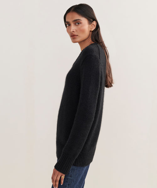 A woman with long dark hair, wearing the black Cashmere Kate V-Neck sweater and blue jeans, stands sideways against a plain light background, looking at the camera with a neutral expression.