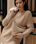 A woman with dark hair wears the Cashmere Kate V-Neck in beige and holds a matching soft handbag. She leans against an indoor railing, looking at the camera with a relaxed expression.