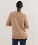 A person with dark hair in a low bun, wearing the Cashmere Kate V-Neck sweater in light brown and blue jeans, stands with their back to the camera against a plain light background.