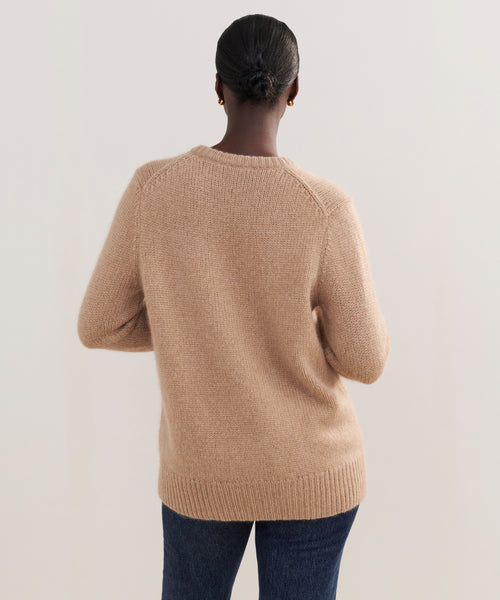 A person with dark hair in a low bun, wearing the Cashmere Kate V-Neck sweater in light brown and blue jeans, stands with their back to the camera against a plain light background.