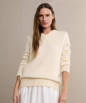 A woman with long brown hair wears the Cashmere Kate V-Neck ivory sweater and a white skirt, standing against a beige background and looking at the camera with a relaxed expression.