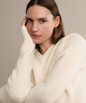 A woman with light brown hair wears the Cashmere Kate V-Neck sweater in ivory, paired with a gold hoop earring, posing serenely against a neutral background with one hand resting on her face.