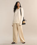 A woman with long dark hair stands against a beige background, wearing the Cashmere Kate V-Neck ivory sweater, loose beige pants, black sandals, and carrying a black and tan woven shoulder bag. She looks relaxed.