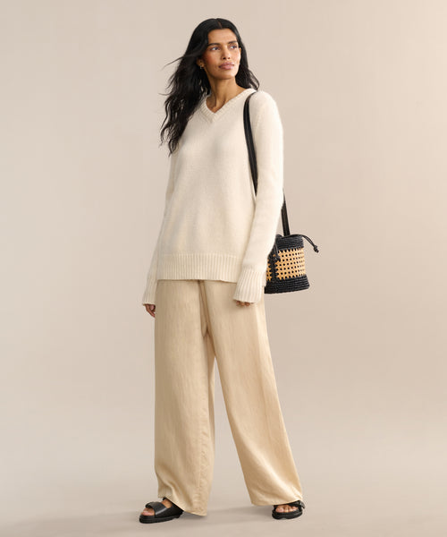 A woman with long dark hair stands against a beige background, wearing the Cashmere Kate V-Neck ivory sweater, loose beige pants, black sandals, and carrying a black and tan woven shoulder bag. She looks relaxed.