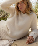 A blonde woman in the Cashmere Kate V-Neck ivory sweater and a white skirt reclines on sandy ground with greenery behind her, shading her eyes with one arm while looking at the camera.