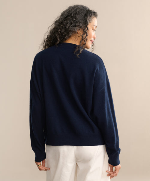A person with long, curly hair faces away, wearing the Cashmere Marina Crewneck in navy blue and white pants against a neutral beige background.