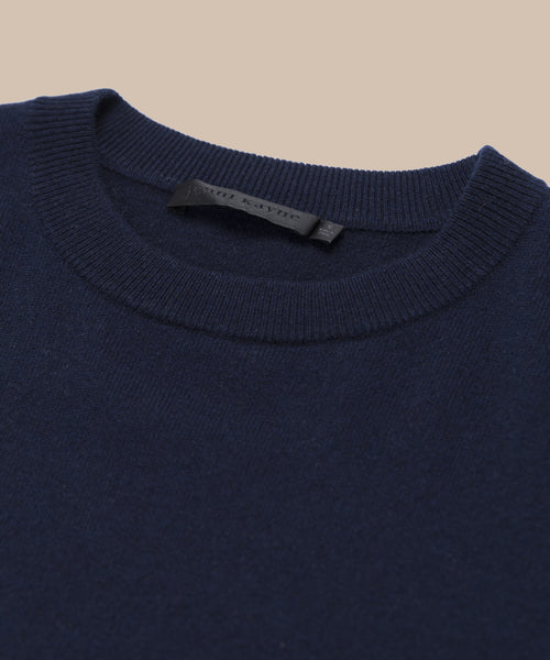 Close-up of the collar and upper section of the dark navy Cashmere Marina Crewneck sweater, featuring a visible black label, set against a neutral beige background.