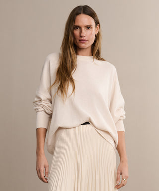 A woman with long brown hair wears the Cashmere Marina Crewneck and a matching pleated skirt, styled in a relaxed silhouette. She stands against a beige background, gazing calmly at the camera.