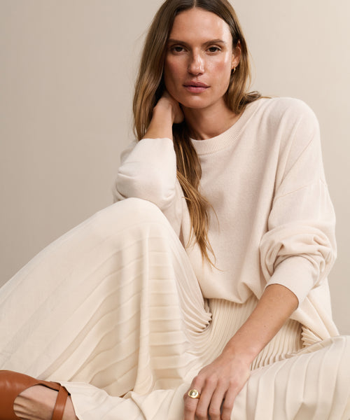 A woman with long, light brown hair sits against a beige background, wearing the oversized Cashmere Marina Crewneck and a pleated cream skirt. She looks relaxed, resting her head on her hand and embodying effortless style.
