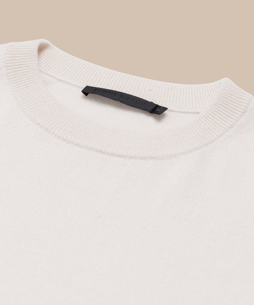 A close-up of the cream-colored Cashmere Marina Crewneck sweater with a ribbed collar and black label sewn into the neckline, shown against a beige background.