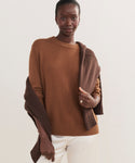 A woman smiles softly at the camera, wearing a camel Cashmere Oversized Crewneck with a brown sweater over her shoulders and off-white pants, standing against a light neutral background.