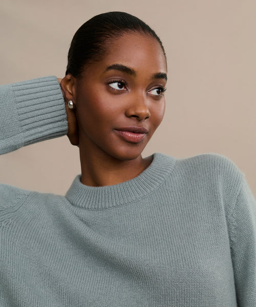 A woman wears a light grayish-blue Cashmere Oversized Crewneck sweater, posing with one hand behind her head against a plain beige background. She has smooth skin, pulled-back hair, and small stud earrings.